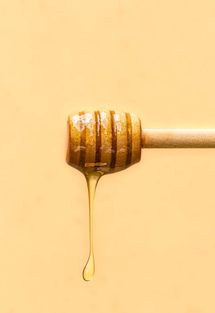 pexels-photo-28714404-1 Close-up of honey drizzling from a wooden dipper against a soft yellow background.